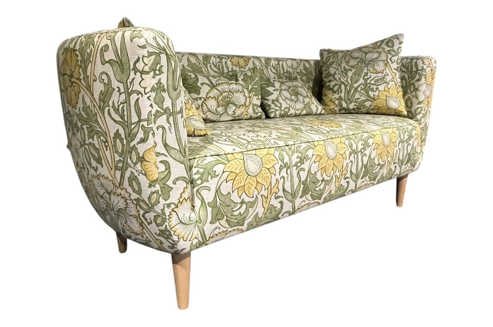 Elegantes Sofa mit geschwungenen Armlehnen und schlanken Holzbeinen. Der Stoffbezug zeigt ein florales Muster mit großen gelben Blumen und grünen Blättern im William-Morris-Stil. Schräg seitliche Ansicht.