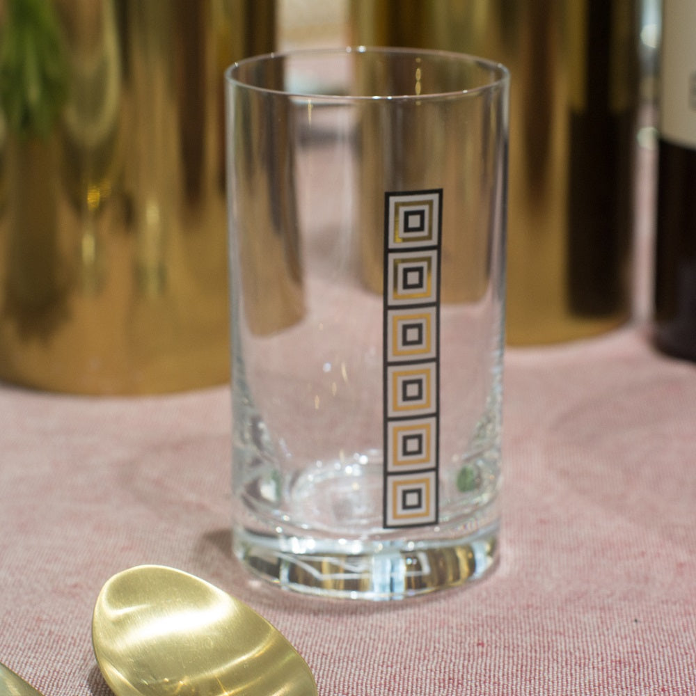 Wasserglas aus der ÖW COLLECTION im zweier Set, Ansicht ganzes glas mit dezenten Aufdruck von schwarzen und goldenen Quadraden, steht auf einem Tisch mit Goldenen Vasen im Hintergrund