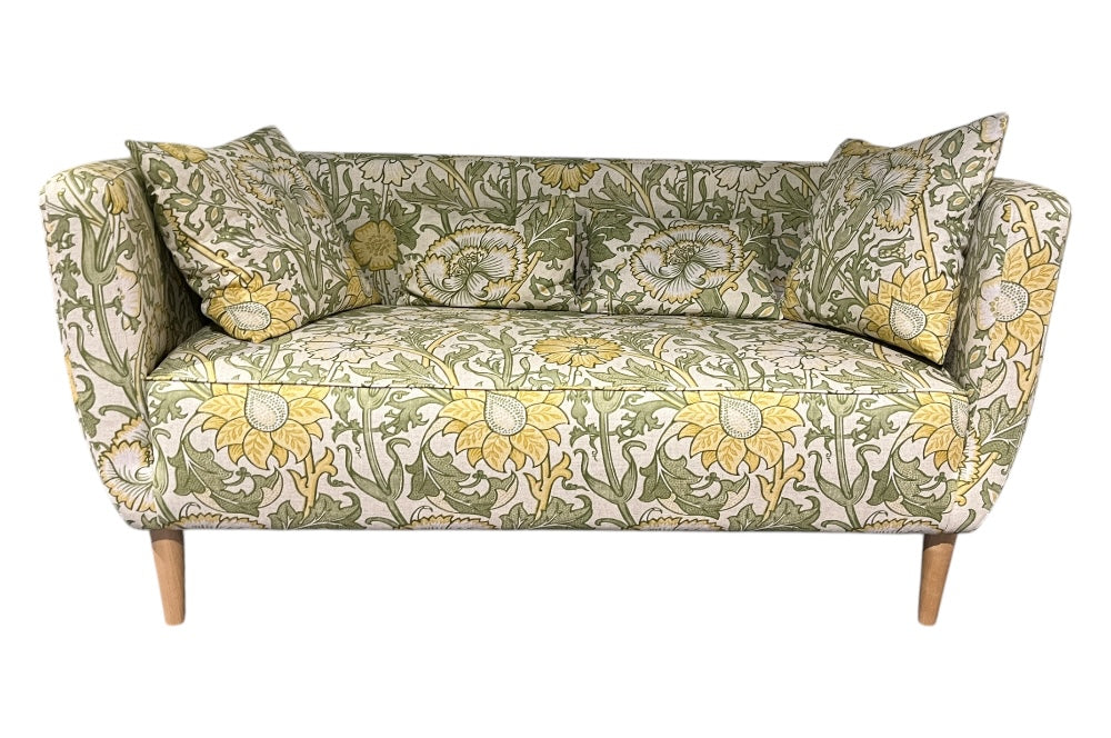 Elegantes Sofa mit geschwungenen Armlehnen und schlanken Holzbeinen. Der Stoffbezug zeigt ein florales Muster mit großen gelben Blumen und grünen Blättern im William-Morris-Stil.